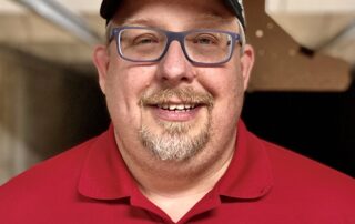 Smiling man wearing glasses and a black cap indoors.